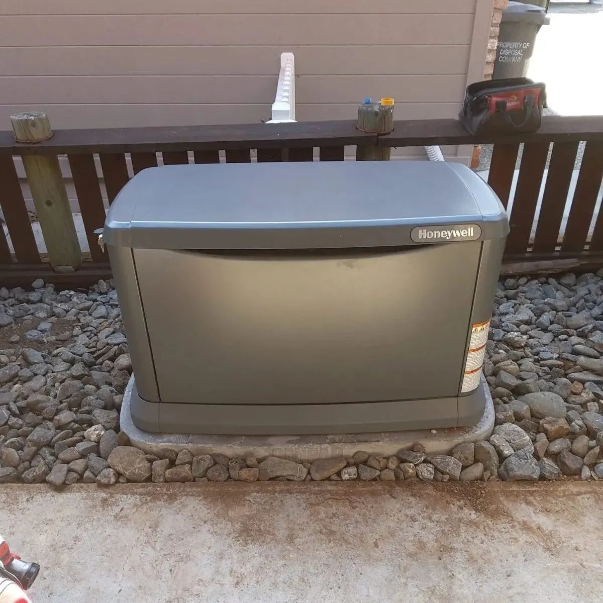 Honeywell standby generator installed on a concrete and gravel base next to a wooden deck railing, with electrical conduit and tools visible nearby.